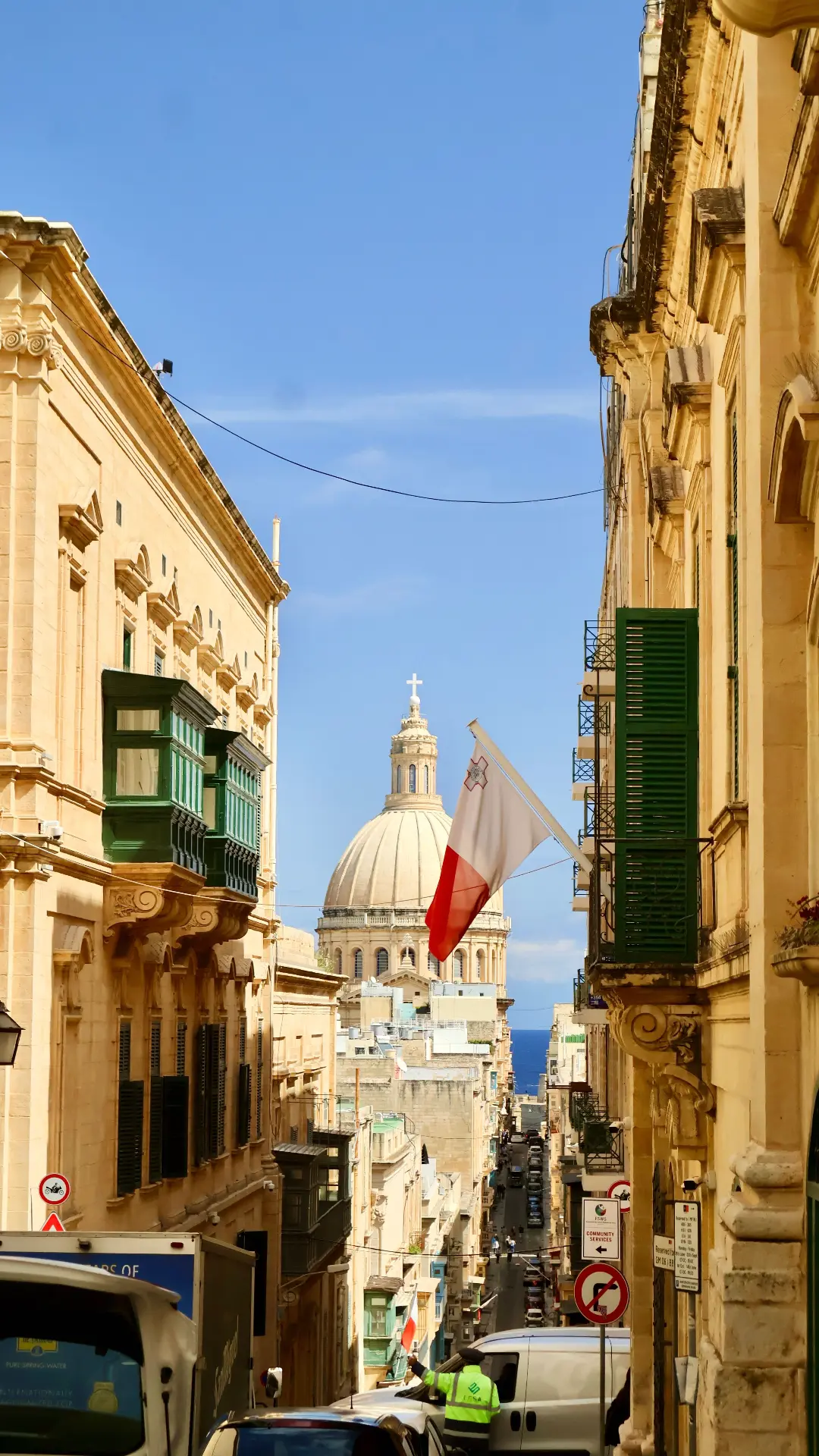 A narrow Valletta street with the Maltese flag and a domed basilica in the distance