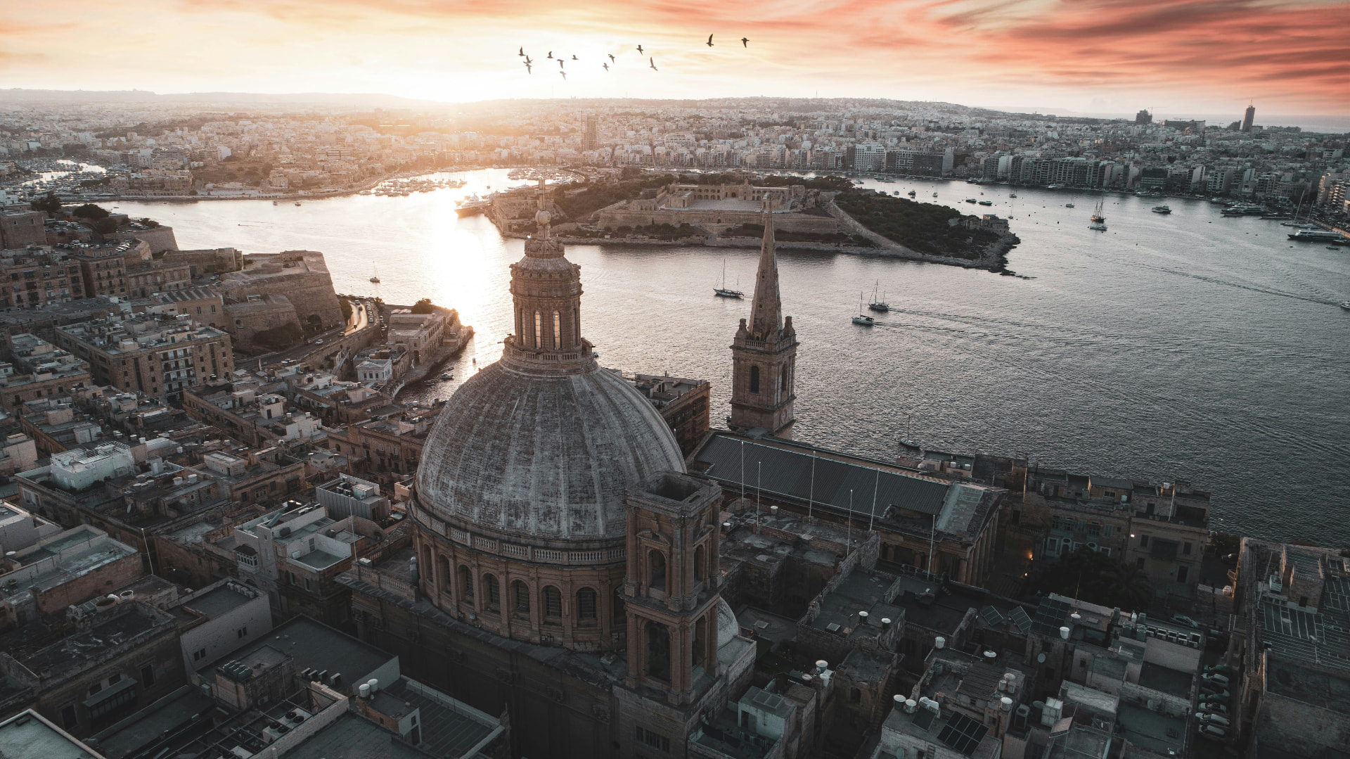 Aerial view of Valletta at sunset, with the basilica dome, spire and Grand Harbour beneath a warm sky